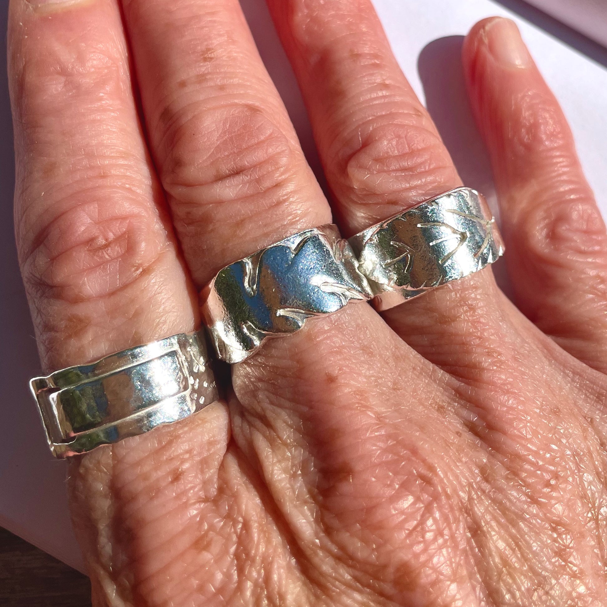 Expanded Skies Wide Ring Workshop Close-up of a hand wearing three silver rings with leaf and abstract designs on a light background