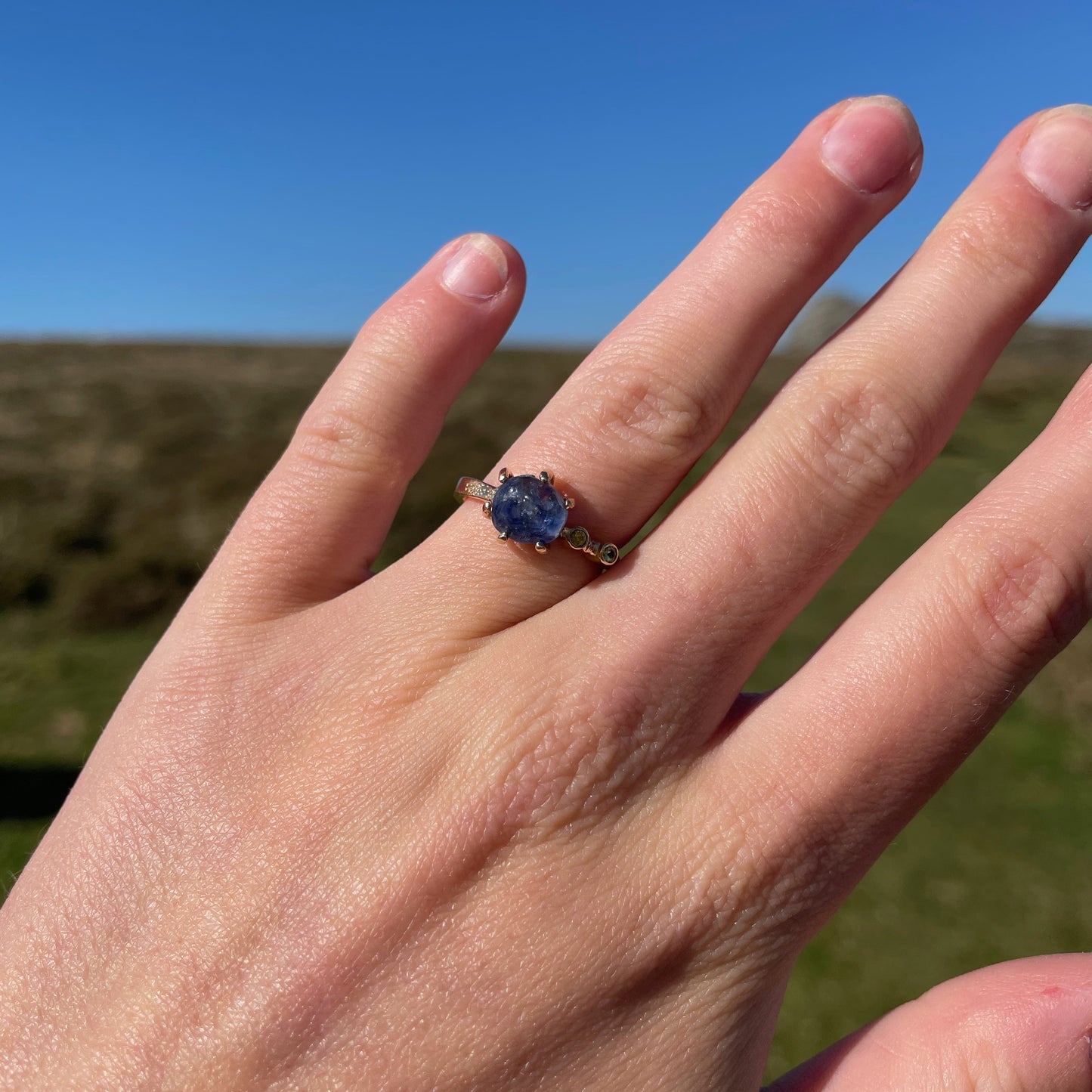 Close up with hand facing he Dartmoor Sun and Skies Sapphire Ring reflects the big Dartmoor skies, the contrasting colours and textures of the moving light over the landscape, created with ethically sourced raw and facetted sapphires and different shades of gold.