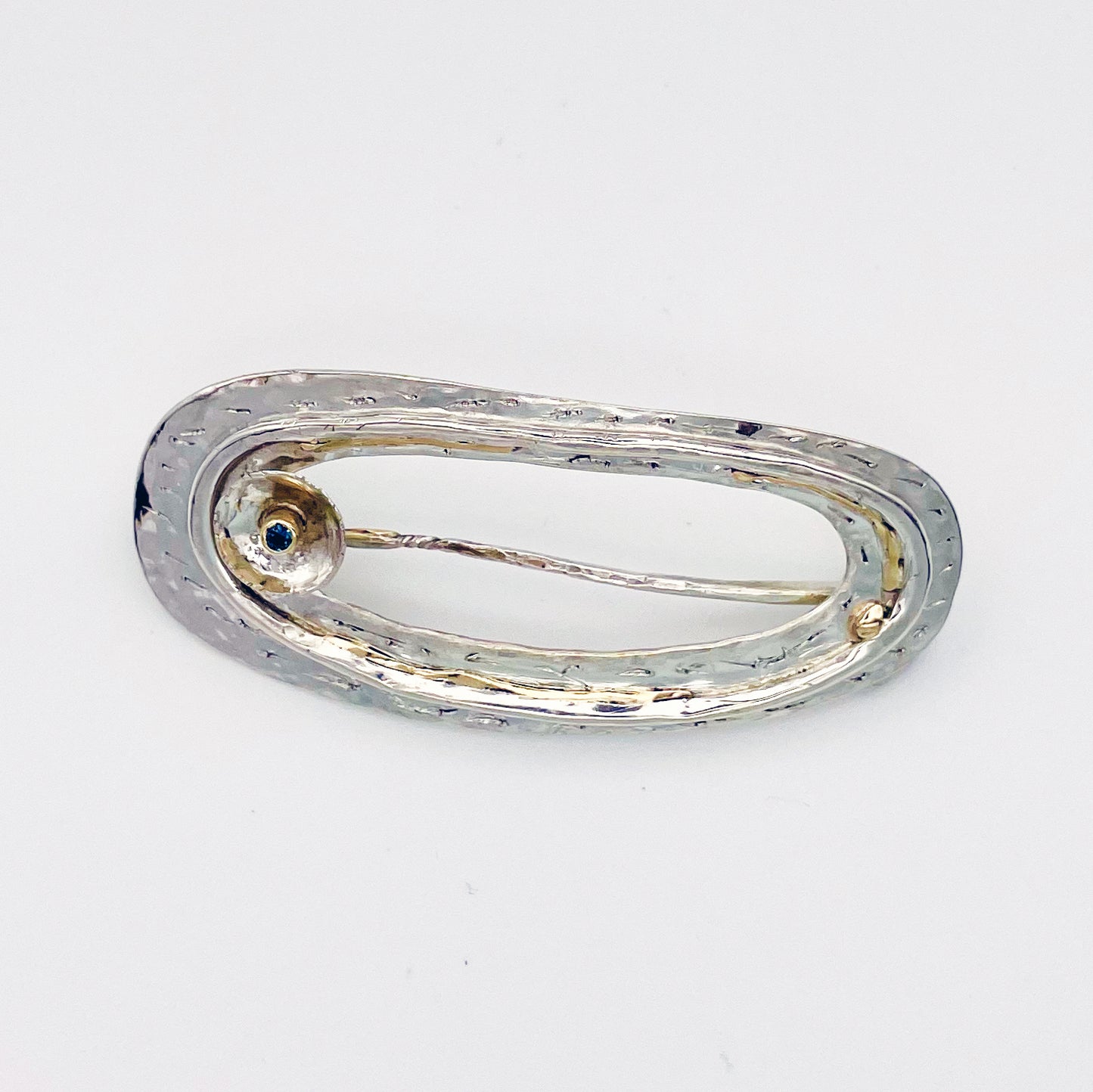 The River Dart at Hembury Brooch is inspired by eddies that glint as they swirl around rocks and tiny islands. The brooch is made from recycled sterling silver that is etched, layered and inlaid with 9ct gold. Totally handcrafted, it has a beautiful 2mm ethically mined teal blue Sri Lankan sapphire set in 9ct recycled gold.