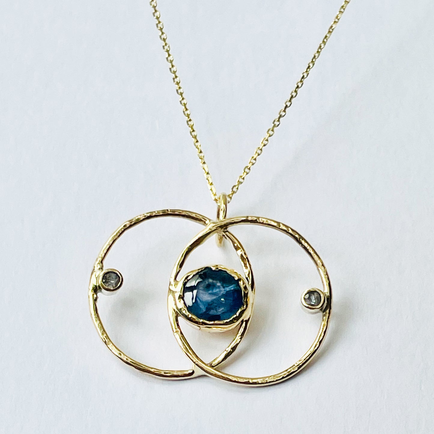 Expanded Skies Blue Sapphire Vesica Piscis Pendant has a large beautiful ethically sourced Sri Lankan blue sapphire with ripples of deep and paler blues, balanced by two faceted white sapphires. The pendent has a handcrafted sculpted and textured setting to reflect the undulating surface of cabochon cut sapphire, in lovely yellow recycled 9ct gold.