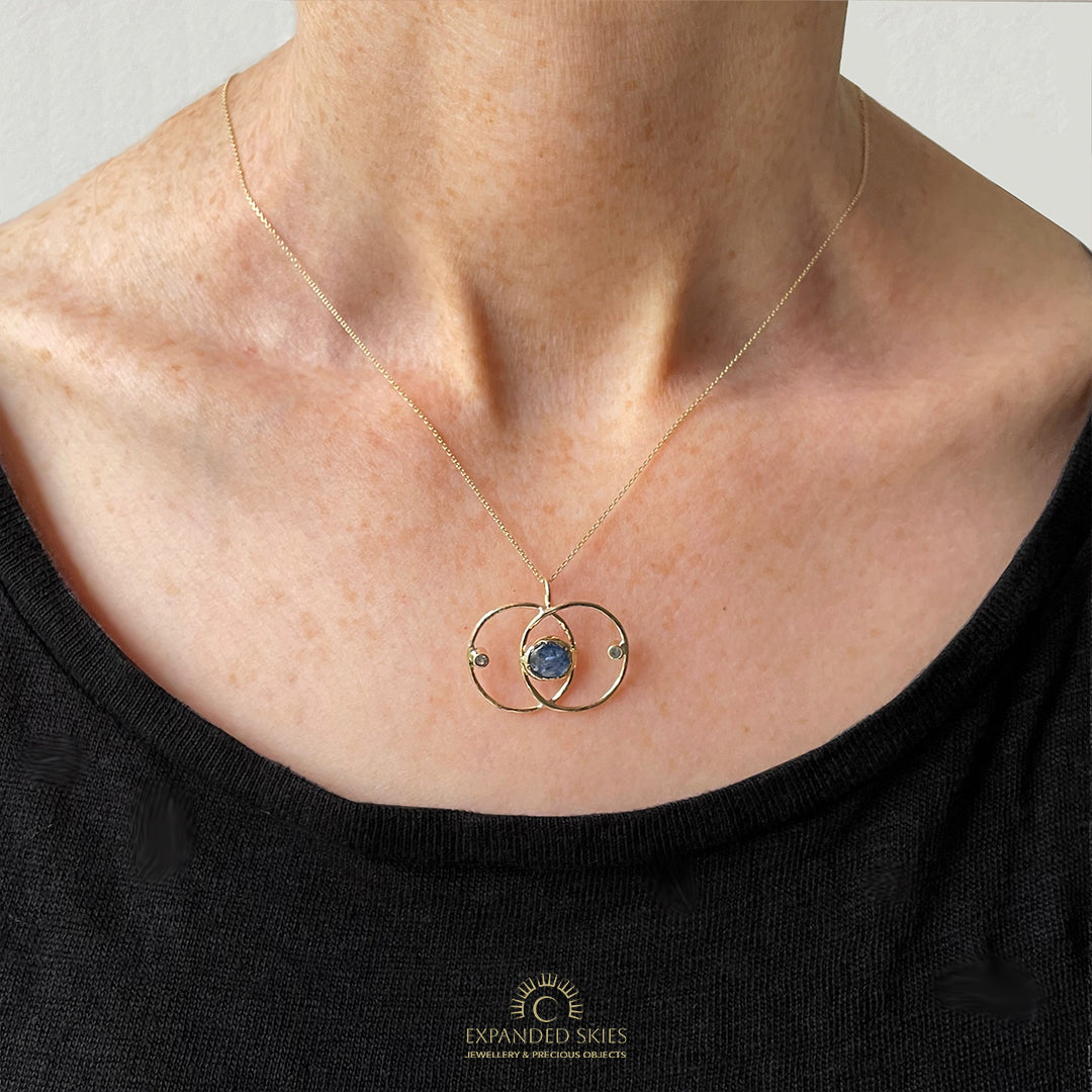 Expanded Skies Blue Sapphire Vesica Piscis Pendant has a large beautiful ethically sourced Sri Lankan blue sapphire with ripples of deep and paler blues, balanced by two faceted white sapphires. The pendent has a handcrafted sculpted and textured setting to reflect the undulating surface of cabochon cut sapphire, in lovely yellow recycled 9ct gold.