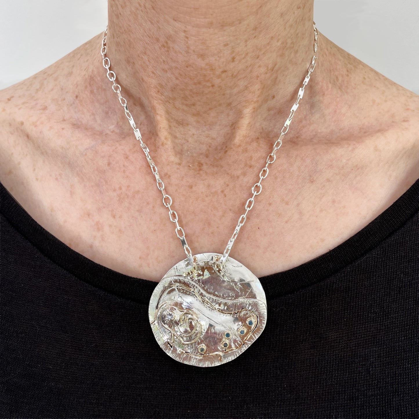 This unique pendant and footpath chain is based on archaeological remains of Hembury Castle, Hill Fort in Hembury Woods, on the edge of Dartmoor. It is created with hand engraving, inlaid with 9ct gold and a beautiful 2mm brilliant cut white Sri Lankan sapphire and five 2mm brilliant cut blue Sri Lankan sapphires ethically sourced from Nineteen48, all set in 9ct gold.