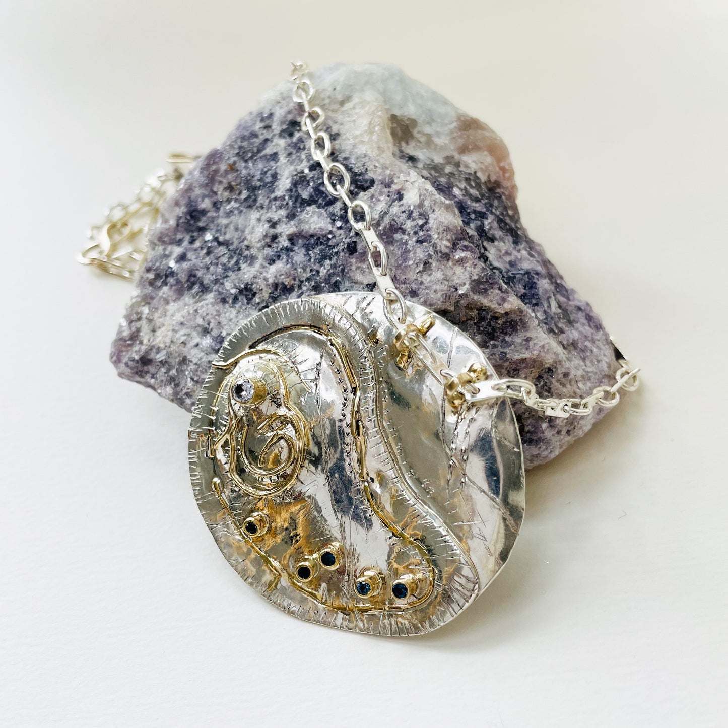 This unique pendant and footpath chain is based on archaeological remains of Hembury Castle, Hill Fort in Hembury Woods, on the edge of Dartmoor. It is created with hand engraving, inlaid with 9ct gold and a beautiful 2mm brilliant cut white Sri Lankan sapphire and five 2mm brilliant cut blue Sri Lankan sapphires ethically sourced from Nineteen48, all set in 9ct gold.