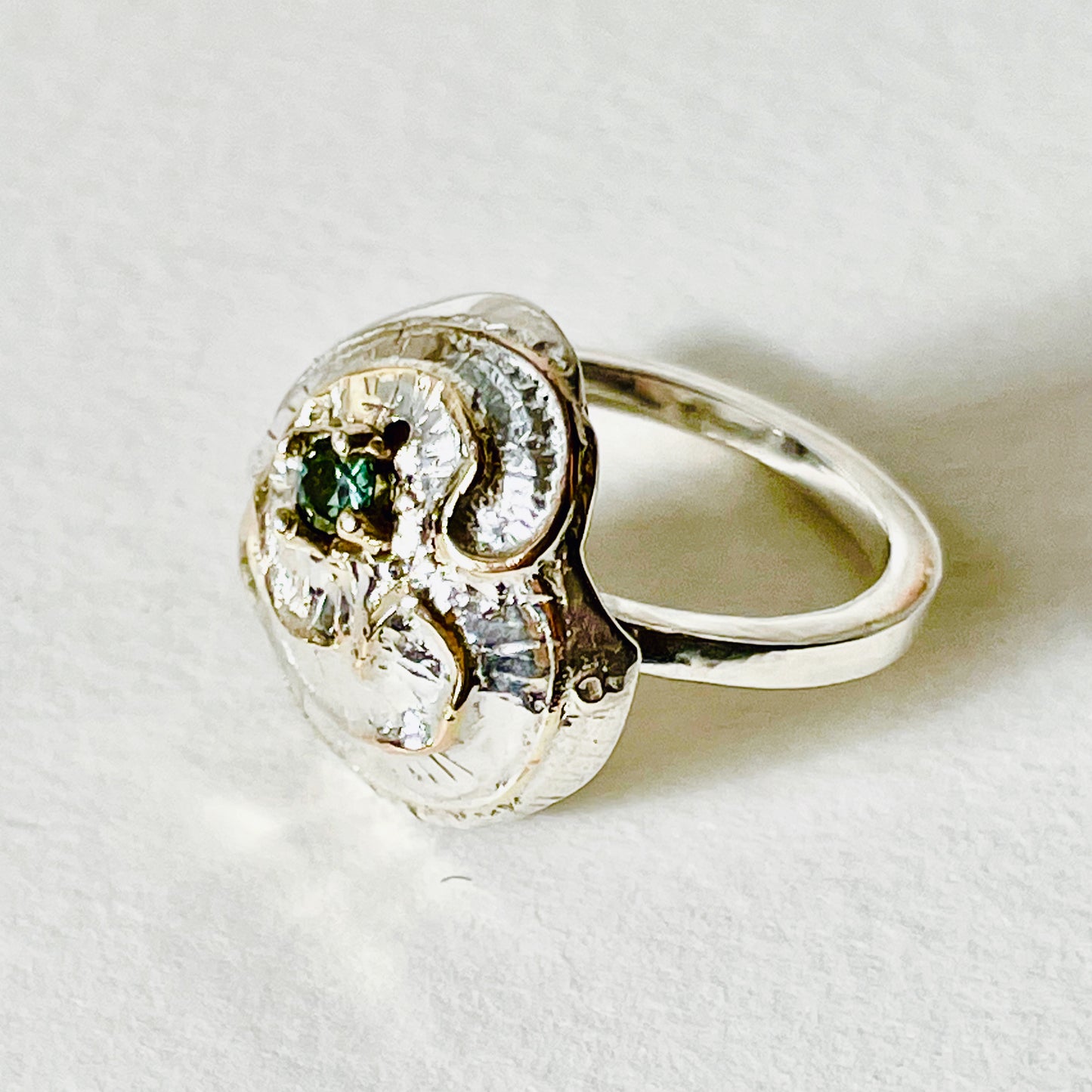 he Hembury Castle Ring design is based on archaeological remains of the mott and bailey Norman castle in Hembury Woods, on the edge of Dartmoor. It is created with hand engraving, inlaid with 9ct gold and set with a beautiful 3mm brilliant cut green Australian sapphire, ethically sourced from Nineteen48. The 'secret' underside of the ring has the symbol of for non-coniferous woodland.