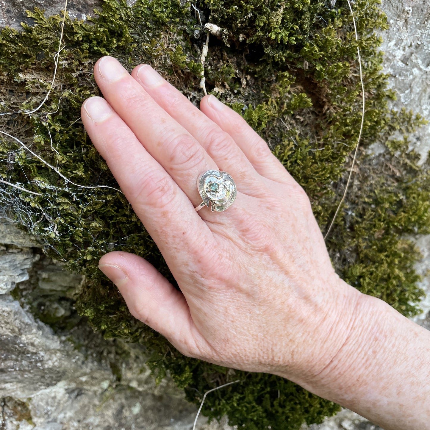 he Hembury Castle Ring design is based on archaeological remains of the mott and bailey Norman castle in Hembury Woods, on the edge of Dartmoor. It is created with hand engraving, inlaid with 9ct gold and set with a beautiful 3mm brilliant cut green Australian sapphire, ethically sourced from Nineteen48. The 'secret' underside of the ring has the symbol of for non-coniferous woodland.