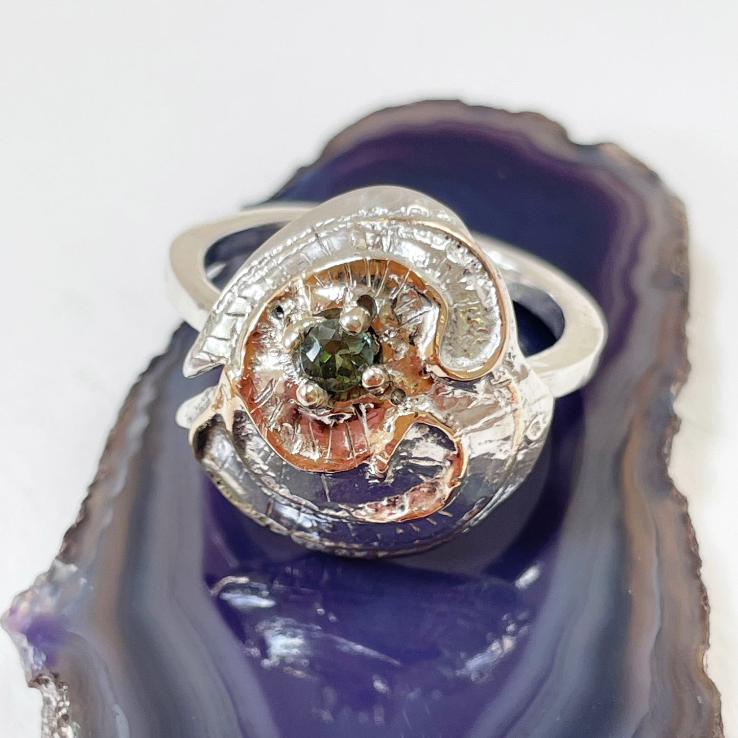 he Hembury Castle Ring design is based on archaeological remains of the mott and bailey Norman castle in Hembury Woods, on the edge of Dartmoor. It is created with hand engraving, inlaid with 9ct gold and set with a beautiful 3mm brilliant cut green Australian sapphire, ethically sourced from Nineteen48. The 'secret' underside of the ring has the symbol of for non-coniferous woodland.
