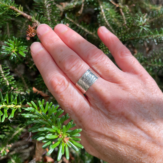 Expanded Skies-Wide Ring Workshop Hand wearing a wide textured silver ring with green foliage in the background