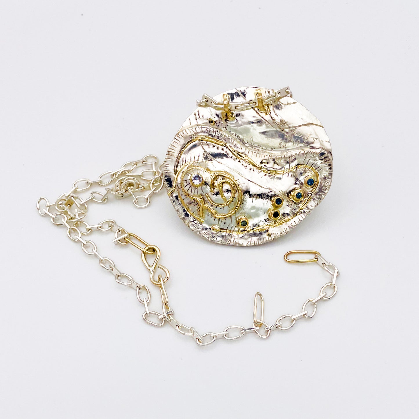 This unique pendant and footpath chain is based on archaeological remains of Hembury Castle, Hill Fort in Hembury Woods, on the edge of Dartmoor. It is created with hand engraving, inlaid with 9ct gold and a beautiful 2mm brilliant cut white Sri Lankan sapphire and five 2mm brilliant cut blue Sri Lankan sapphires ethically sourced from Nineteen48, all set in 9ct gold.