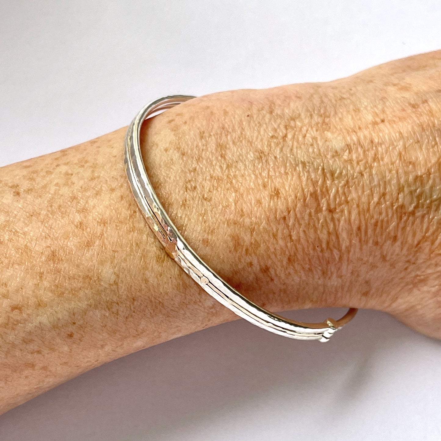 The Hembury Woods Hinged Bangle evokes the river Dart River at Hembury, where the water eddies and glints as it swirls around the rocks. Silver is formed to reflect the undulating river course and the glinting water, inlaid with 9ct gold. The bangle is totally handcrafted on the edge of Dartmoor and made from high quality recycled sterling silver, and recycled 9ct gold and with a textured hammered finish that reflects the light. Understated and beautiful, it can be worn every day. It can be personalised.