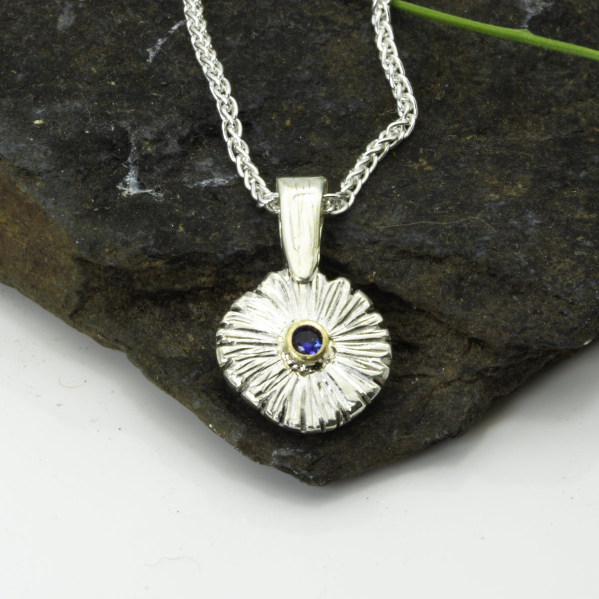 The Expanded Skies Dartmoor Daisy Blue Sapphire Necklace in sterling silver and 9ct gold is inspired by the Erigeron Karvinskianus, commonly known as the 'Dartmoor Daisy'. It comes with 16” Italian spiga rolled chain, made from recycled sterling silver. The daisy pendant measures 12mm, the bail measures 6mm, total length 18mm with a depth of 2mm (approx). Each piece is individually saw pierced, hand engraved and
unique. The ethically sourced 2.5mm is a beautiful brilliant blue, facetted Sri Lankan sapphire,