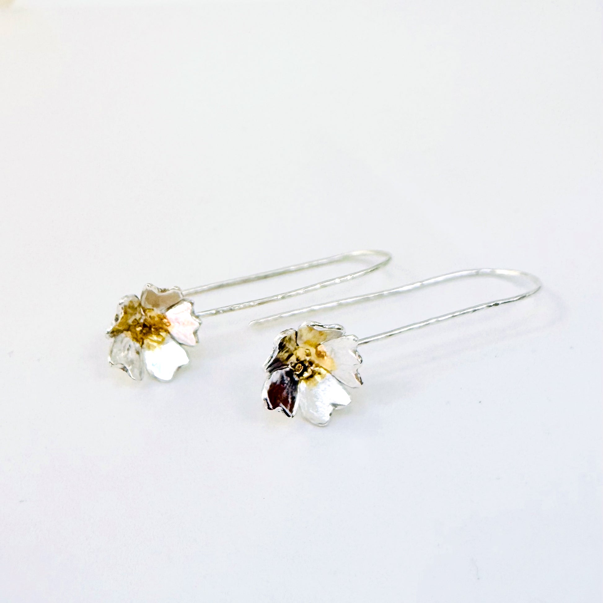 The Expanded Skies Dartmoor Field Rose Drop Earrings capture the joyous simple beauty of the field rose, Rosa arvensis, that is seen weaving brightly through the Dartmoor hedgerows and woodland. Hand pierced and formed in recycled fine silver with recycled 24ct gold accents and recycled sterling silver ear wires. 