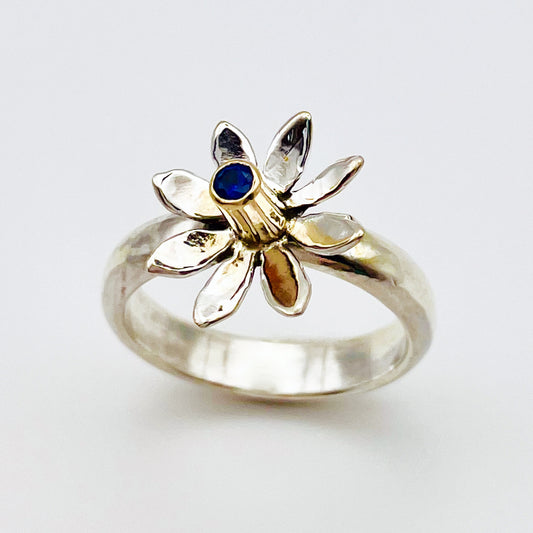 The Expanded Skies Breeze Blown Daisy Sapphire Ring is joyful! A beautiful brilliant bright blue, ethically sourced, Sri Lankan sapphire is set in solid 9ct gold that reflects onto to the high polish recycled silver petals, giving a sense of movement. The ring is totally handcrafted on the edge of Dartmoor.