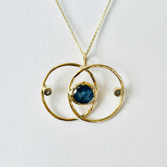 Expanded Skies Blue Sapphire Vesica Piscis Pendant has a large beautiful ethically sourced Sri Lankan blue sapphire with ripples of deep and paler blues, balanced by two faceted white sapphires. The pendent has a handcrafted sculpted and textured setting to reflect the undulating surface of cabochon cut sapphire, in lovely yellow recycled 9ct gold.