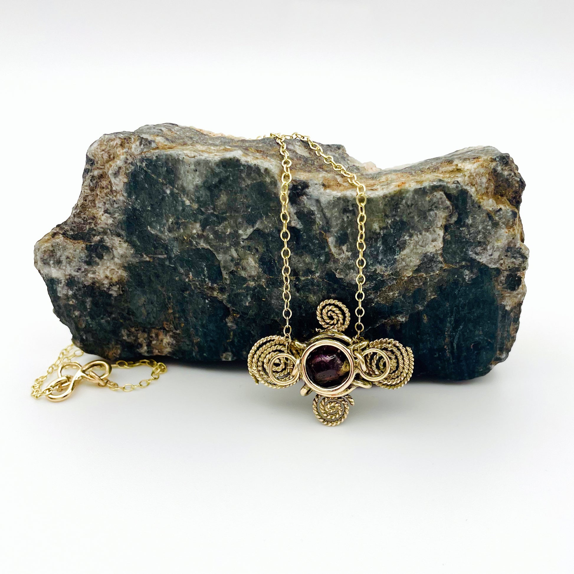 The Expanded Skies Raw Garnet Spiral Filigree Necklace has a beautiful raw Norwegian garnet set in 9ct gold. The garnet is set in a distinctive completely handcrafted 9ct gold filigree and claw setting. It features the neolithic symbol of the spiral, representing the life force and the cycles of life. This is a unique piece, one-of-a-kind. Handcrafted on the edge of Dartmoor and made from high quality recycled gold, Hallmarked by the London Assay Office.