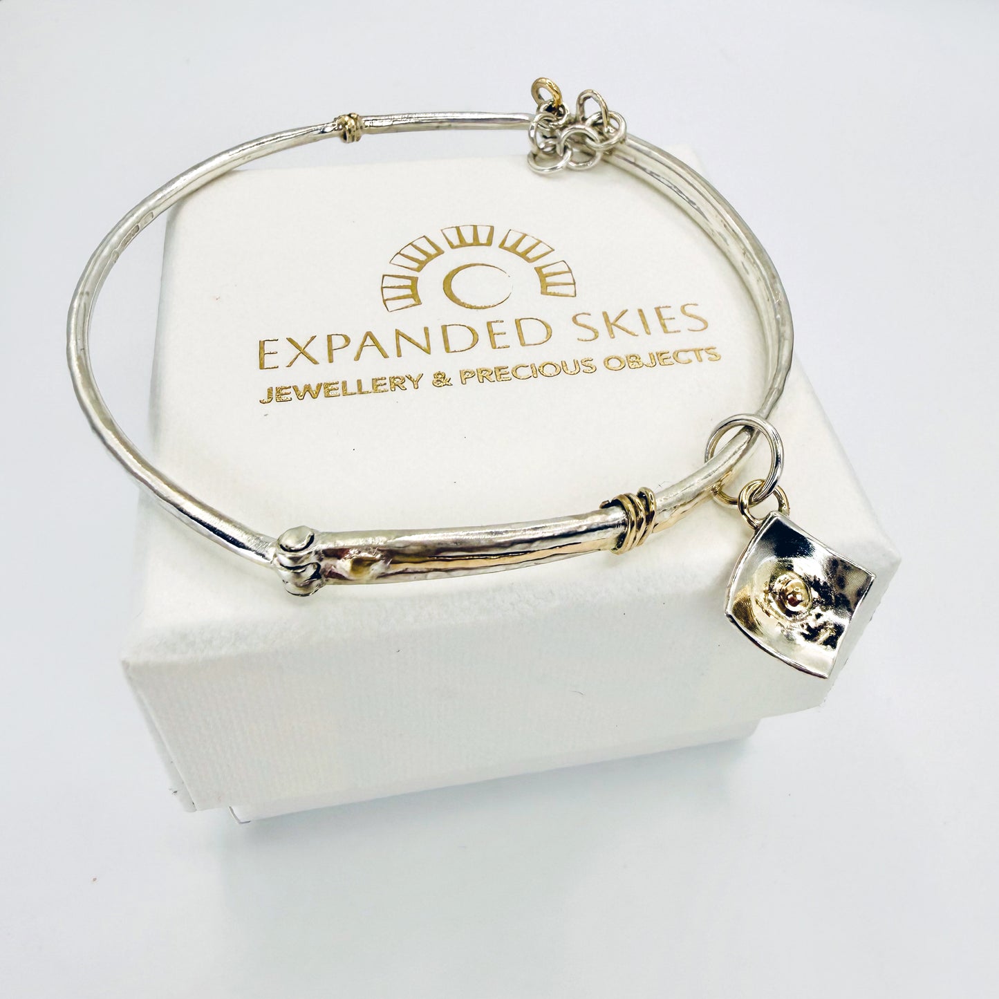 The River Dart at Hembury Hinged Bangle evokes the River Dart that flows through Hembury Woods, where the water eddies and glints as it swirls around the rocks. 
The bangle is totally handcrafted on the edge of Dartmoor and made from high quality recycled sterling silver, inlaid with recycled 9ct gold, with a textured finish that reflects the light. Shown with Expanded Skies box handcrafted in Cornwall.