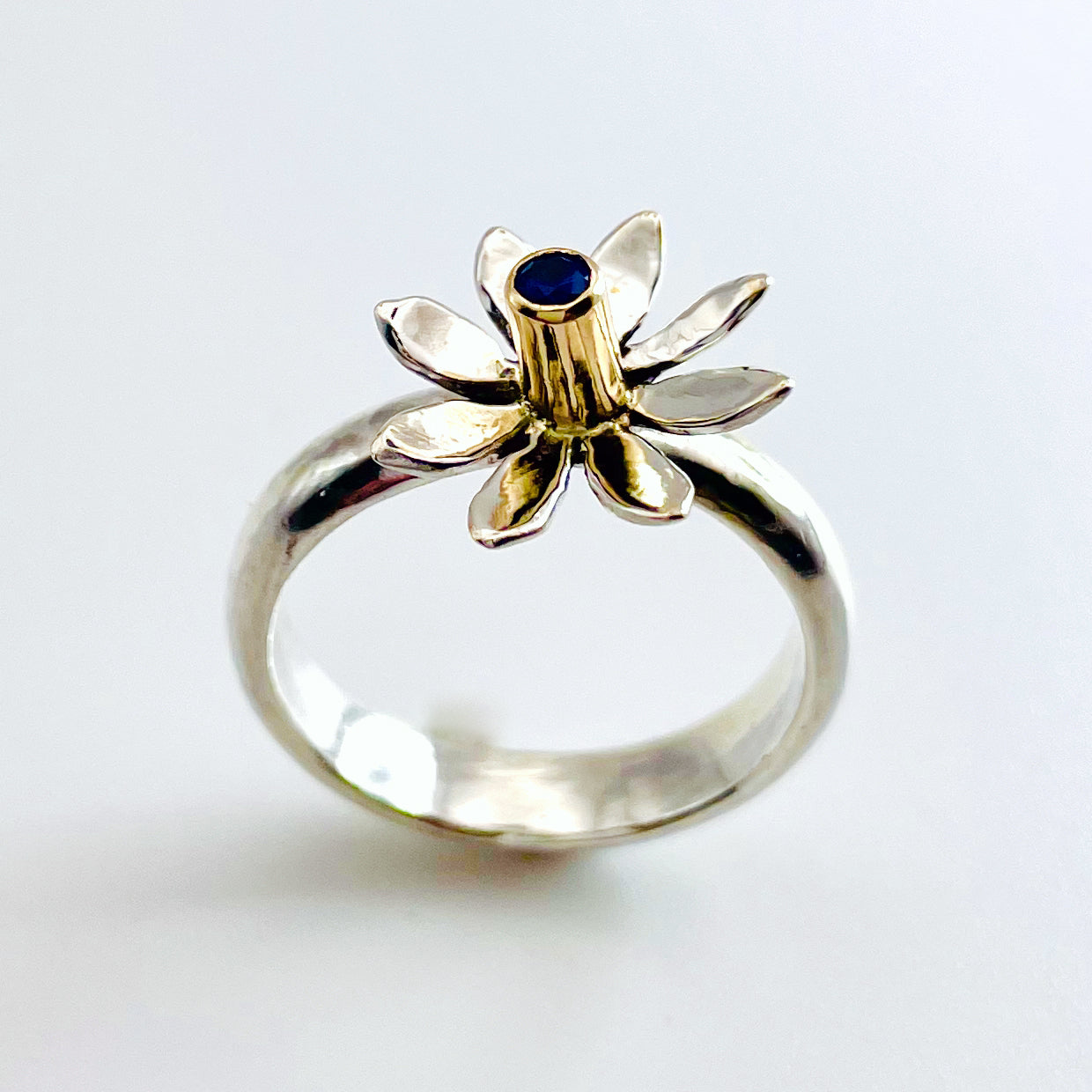 The Expanded Skies Breeze Blown Daisy Sapphire Ring is joyful! A beautiful brilliant bright blue, ethically sourced, Sri Lankan sapphire is set in solid 9ct gold that reflects onto to the high polish recycled silver petals, giving a sense of movement. The ring is totally handcrafted on the edge of Dartmoor.