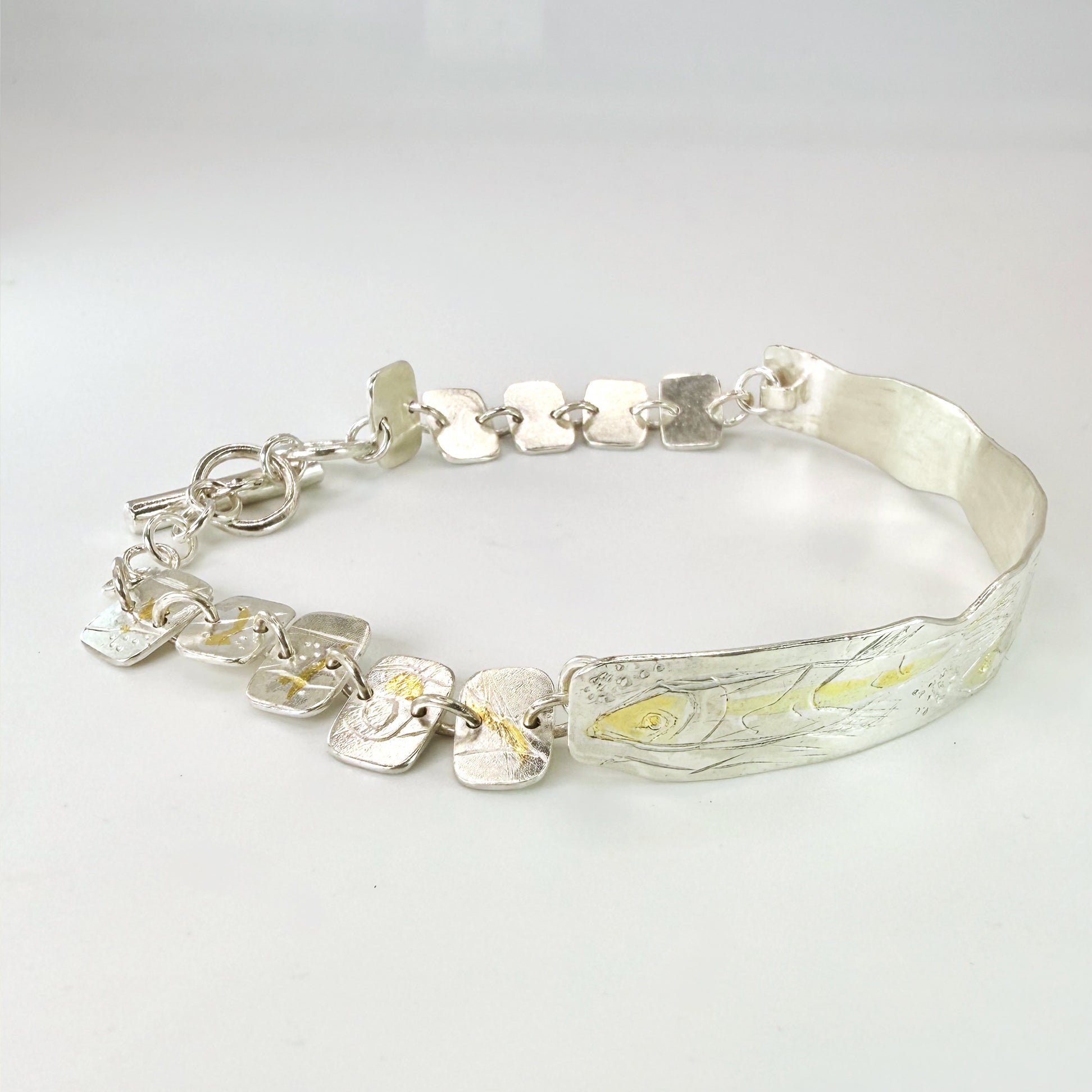 The Expanded Skies Dartmoor minnows bracelet captures the graceful beauty of the minnows as they somersault through the glistening River Dart at Hembury Woods, created in fine and sterling silver with 24ct gold accents. The chain is created with hand engraved fine silver panels with 24ct gold accents depict the moving river. Understated and beautiful, it can be worn every day or for special occasions, for men and women.The bracelet is totally handcrafted on the edge of Dartmoor from recycled precious metals
