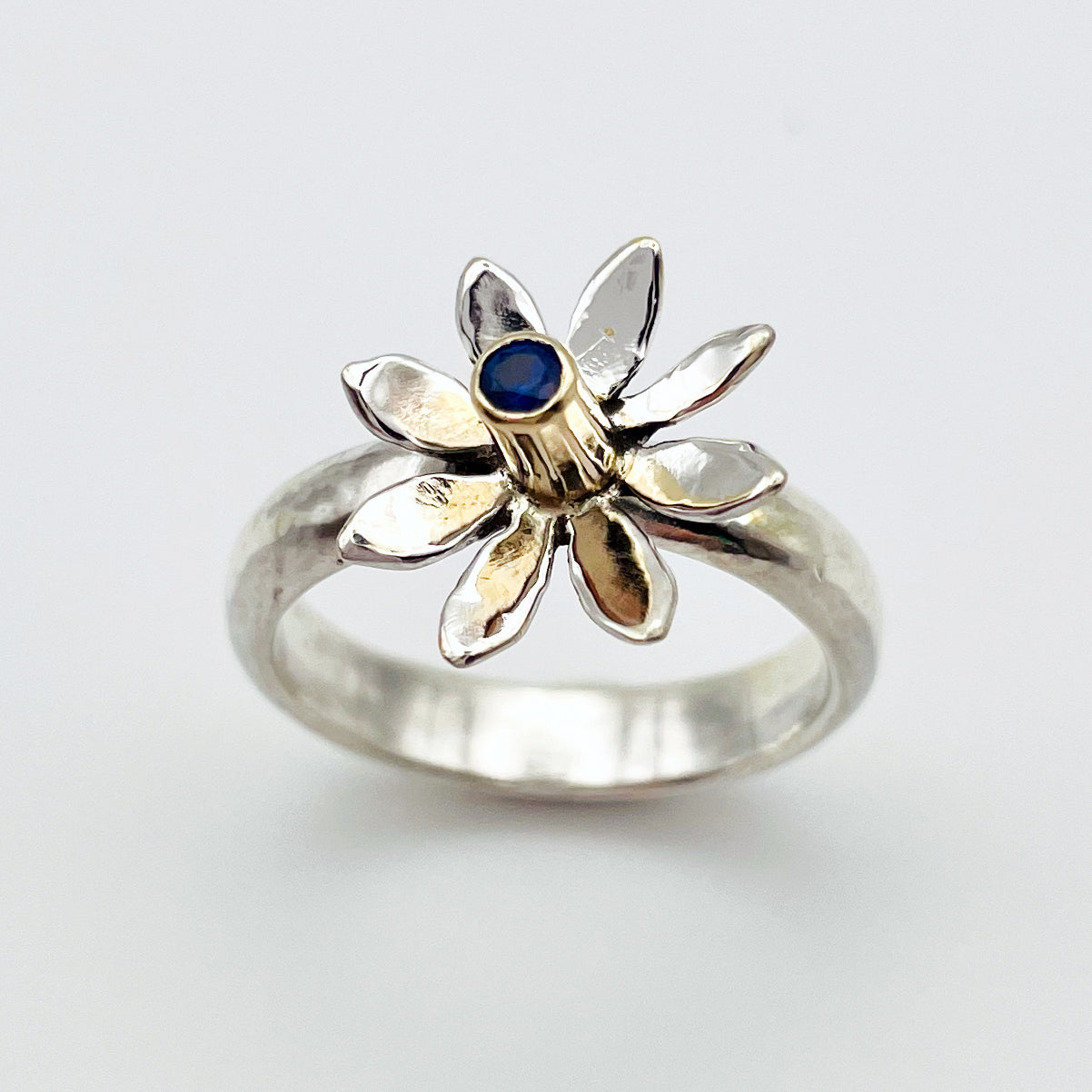 The Expanded Skies Breeze Blown Daisy Sapphire Ring is joyful! A beautiful brilliant bright blue, ethically sourced, Sri Lankan sapphire is set in solid 9ct gold that reflects onto to the high polish recycled silver petals, giving a sense of movement. The ring is totally handcrafted on the edge of Dartmoor.