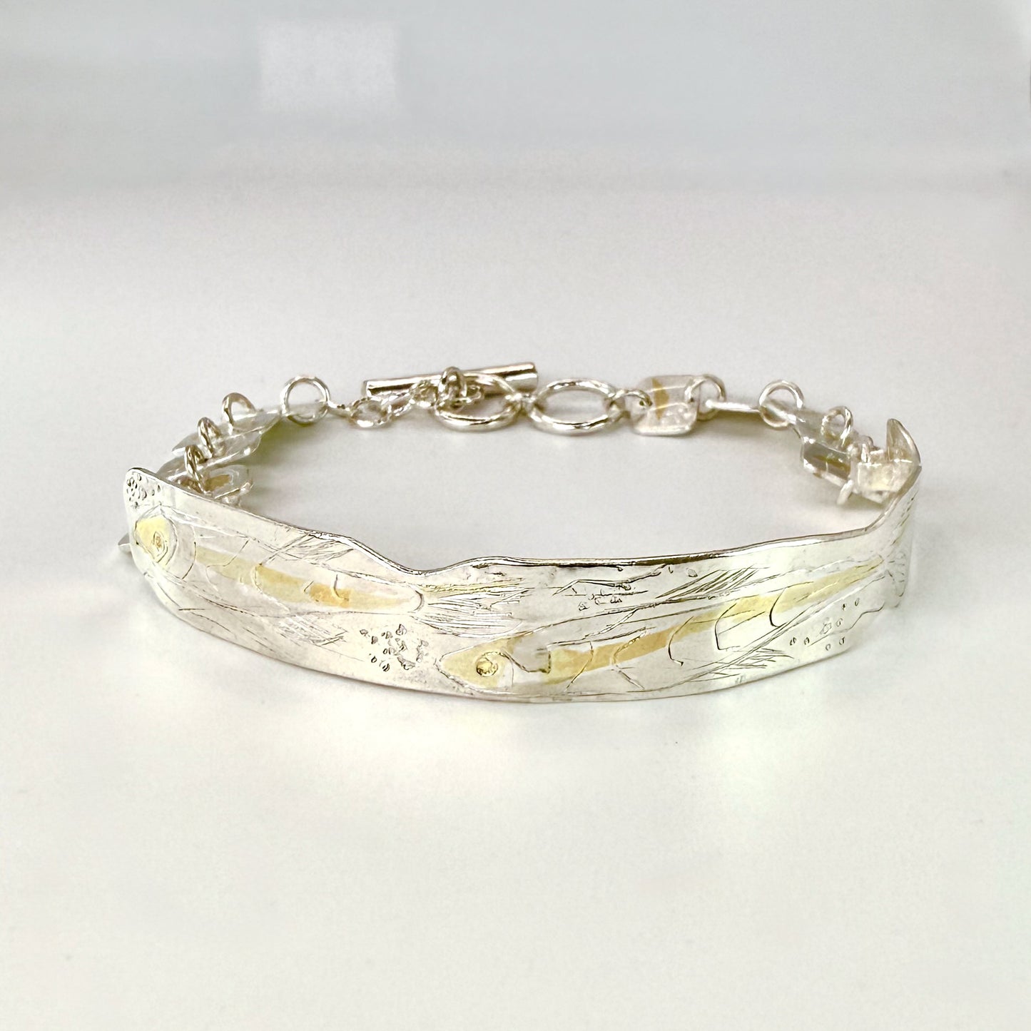 The Expanded Skies Dartmoor minnows bracelet captures the graceful beauty of the minnows as they somersault through the glistening River Dart at Hembury Woods, created in fine and sterling silver with 24ct gold accents. The chain is created with hand engraved fine silver panels with 24ct gold accents depict the moving river. Understated and beautiful, it can be worn every day or for special occasions, for men and women.The bracelet is totally handcrafted on the edge of Dartmoor from recycled precious metals