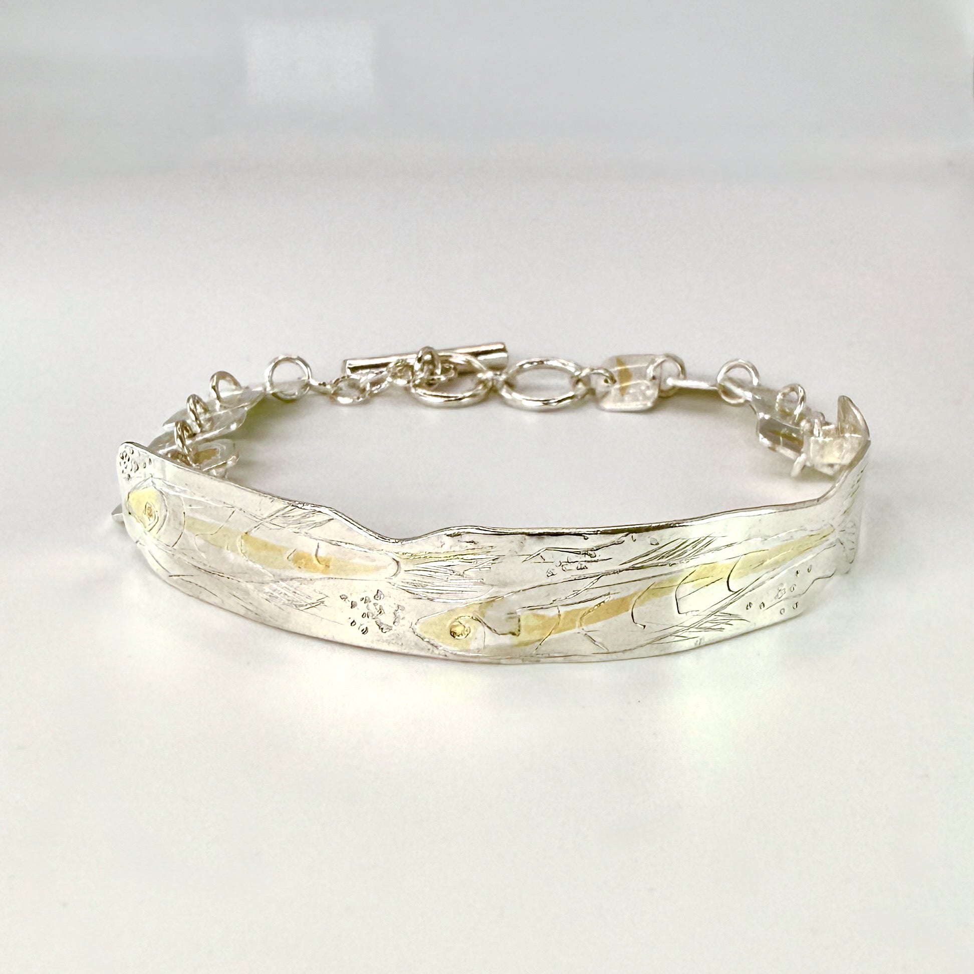 The Expanded Skies Dartmoor minnows bracelet captures the graceful beauty of the minnows as they somersault through the glistening River Dart at Hembury Woods, created in fine and sterling silver with 24ct gold accents. The chain is created with hand engraved fine silver panels with 24ct gold accents depict the moving river. Understated and beautiful, it can be worn every day or for special occasions, for men and women.The bracelet is totally handcrafted on the edge of Dartmoor from recycled precious metals