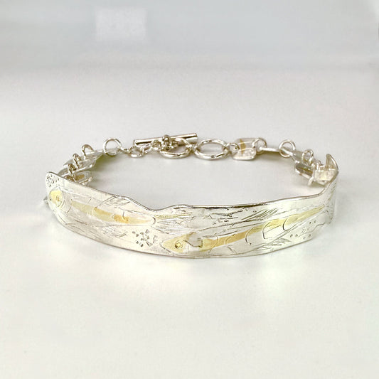 The Expanded Skies Dartmoor minnows bracelet captures the graceful beauty of the minnows as they somersault through the glistening River Dart at Hembury Woods, created in fine and sterling silver with 24ct gold accents. The chain is created with hand engraved fine silver panels with 24ct gold accents depict the moving river. Understated and beautiful, it can be worn every day or for special occasions, for men and women.The bracelet is totally handcrafted on the edge of Dartmoor from recycled precious metals