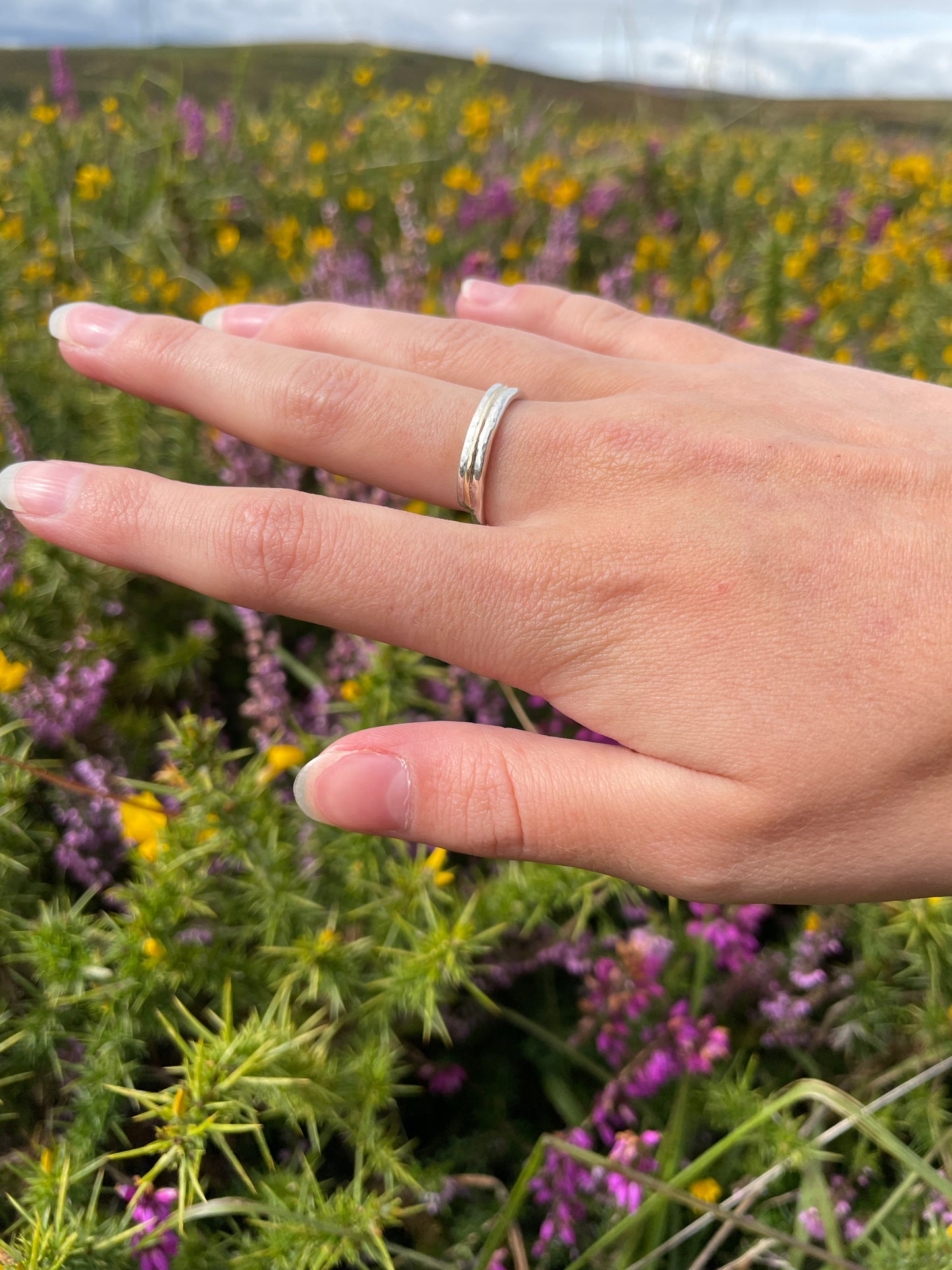 Woman wearing The Dartmoor Dart Valley Hammered Ring, inlaid with 9ct Gold, is inspired by the River Dart, that viewed from high on top of granite moorland, glints and snakes through the steep sides of the deeply wooded valley.  The hammered band is handcrafted from a single band of sold sterling recycled silver at back that flows into a recycled 9ct gold band at the front, flanked by two silver bands.  Wear everyday, perfect as a wedding ring or to accompany any outfit, understated, beautiful.