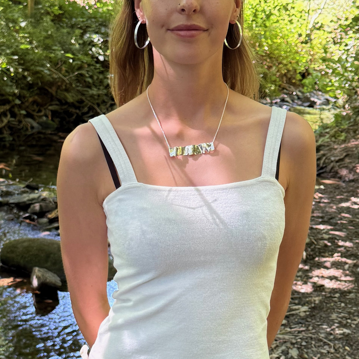 Expanded Skies River Teign Necklace is inspired by the River Teign as it moves around rocks in the Gorge, drawn with hand engraving and 24ct gold accents. Worn by a woman.