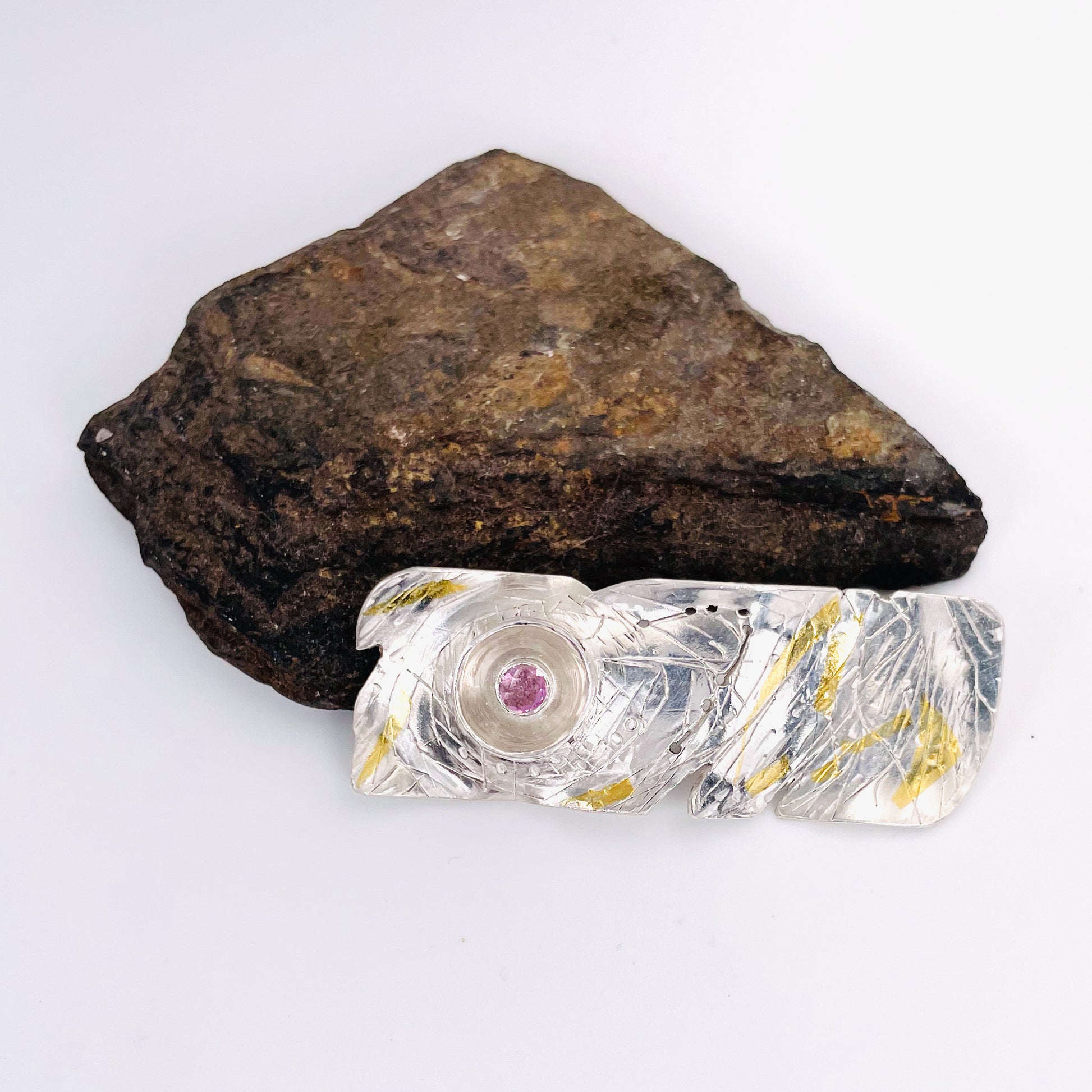 The Expanded Skies Expanded Skies Dartmoor Sun and Stars Pink Sapphire Brooch is inspired by the rising sun and the emerging stars in Dartmoor skies. Created with hand engraving and gold accents with a beautiful pink sapphire.