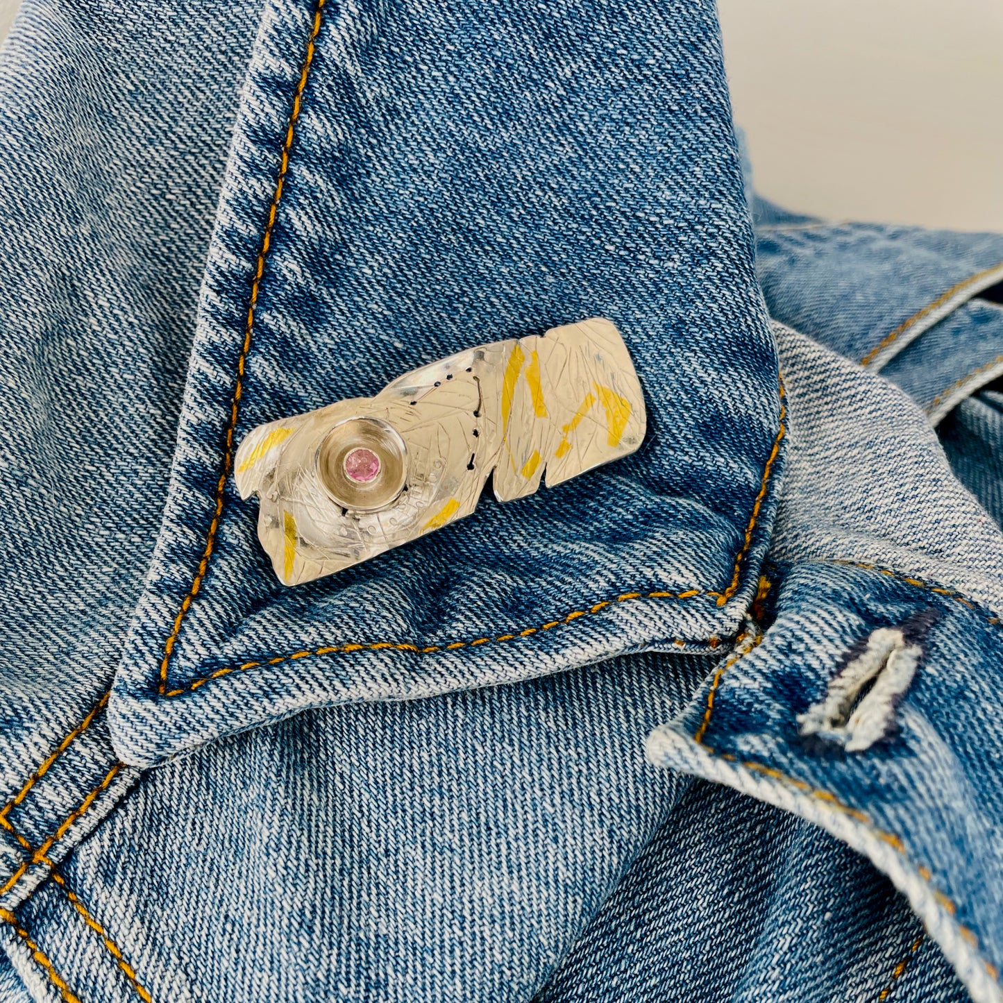 The Expanded Skies Expanded Skies Dartmoor Sun and Stars Pink Sapphire Brooch is inspired by the rising sun and the emerging stars in Dartmoor skies. Created with hand engraving and gold accents with a beautiful pink sapphire. Pined to denim jacket.
