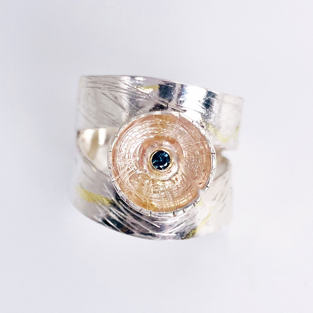 Expanded Skies River Teign Ring captures the sculptural forms of the River Teign as it moves around rocks in the Gorge, drawn with hand engraving and set with a twinkling brilliant blue sapphire in 9ct gold. The hand engraving has gold accents created by fusing 24ct gold to the silver using the kaum boo gilding process.