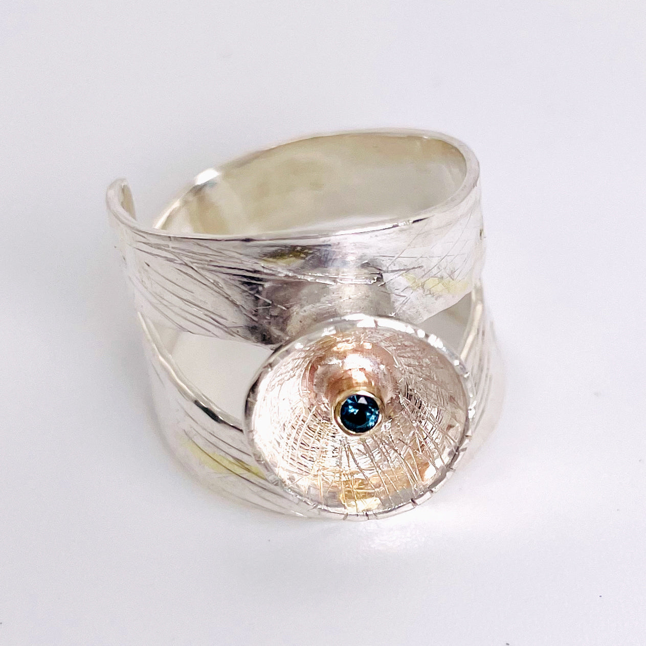 Expanded Skies River Teign Ring captures the sculptural forms of the River Teign as it moves around rocks in the Gorge, drawn with hand engraving and set with a twinkling brilliant blue sapphire in 9ct gold. The hand engraving has gold accents created by fusing 24ct gold to the silver using the kaum boo gilding process.