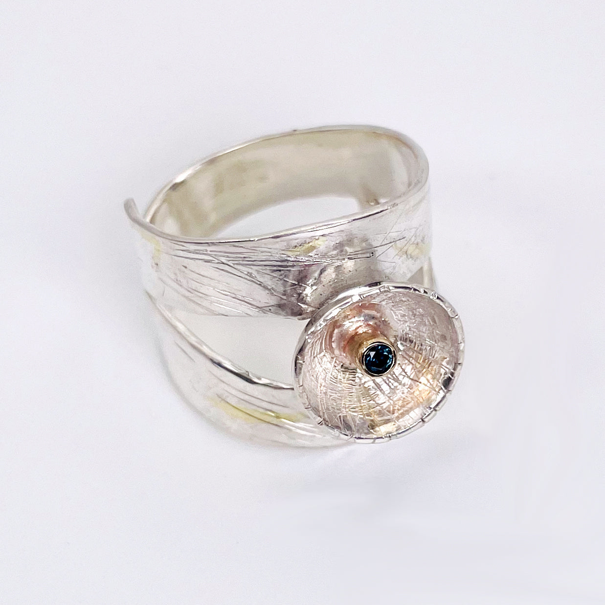 Expanded Skies River Teign Ring captures the sculptural forms of the River Teign as it moves around rocks in the Gorge, drawn with hand engraving and set with a twinkling brilliant blue sapphire in 9ct gold. The hand engraving has gold accents created by fusing 24ct gold to the silver using the kaum boo gilding process.